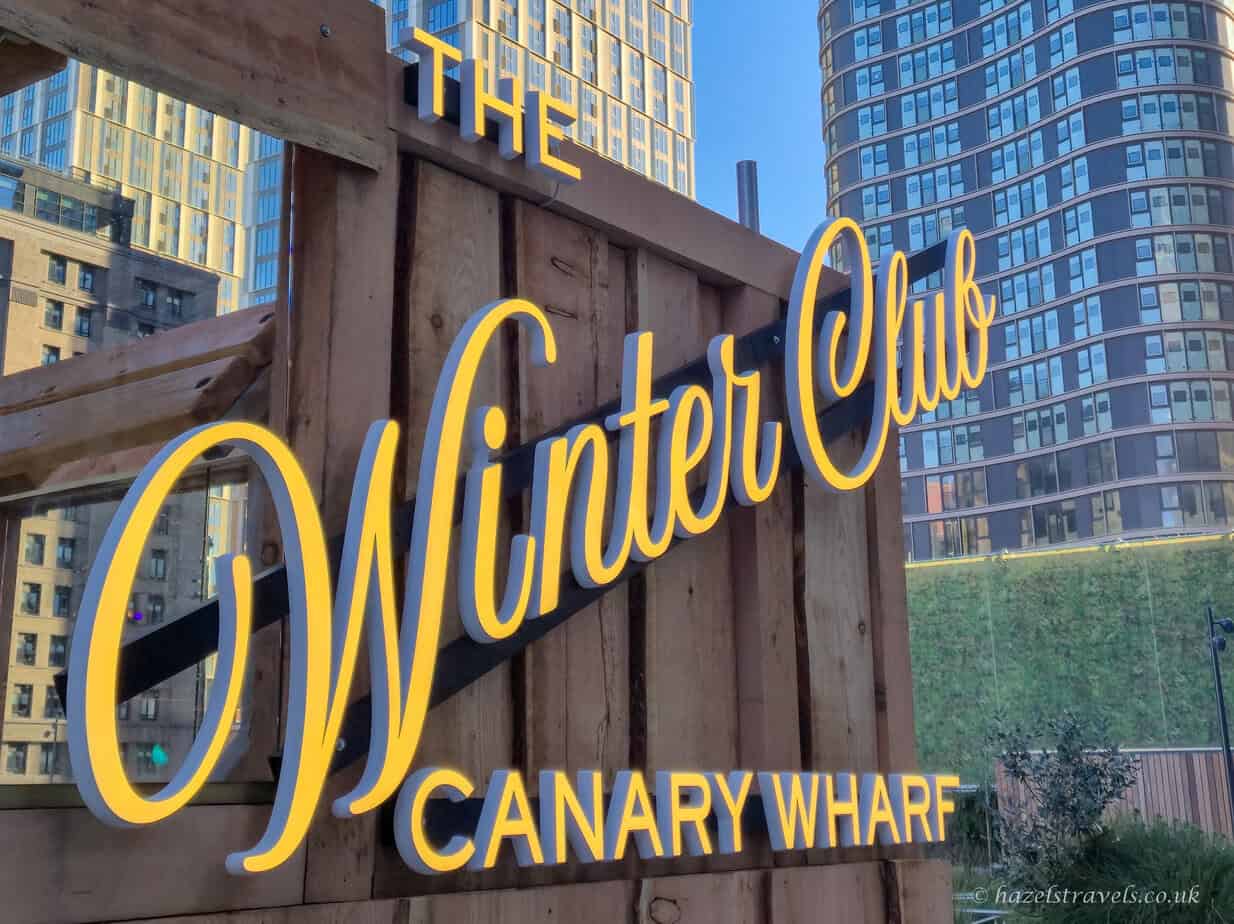 The Winter Club Canary Wharf sign in bold yellow letters on a wooden backdrop, with tall glass buildings rising behind under a clear blue sky.