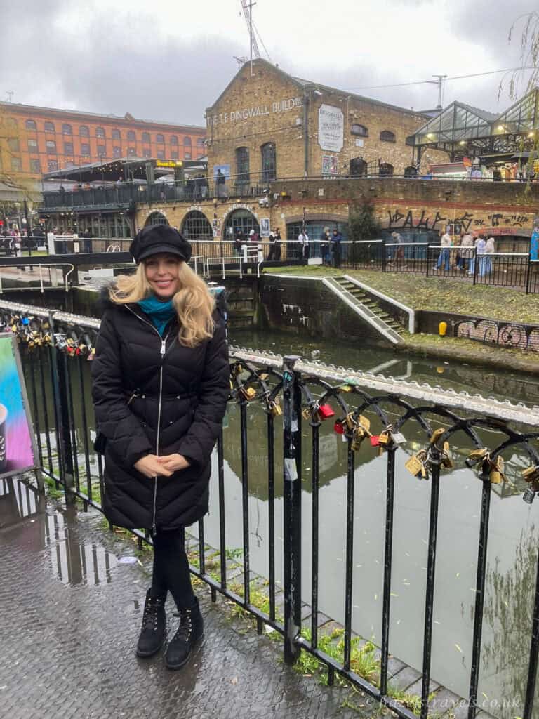 Woman in a black coat and hat standing by the canal at Camden Market in London, with colourful buildings, railings covered in love locks, and waterside walkways in the background.