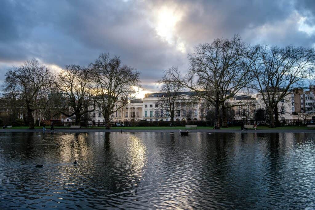 Winter view of Regent’s Park in London with a calm lake reflecting a cloudy blue and grey sky, bare trees lining the water, and pale cream Regency buildings in the background