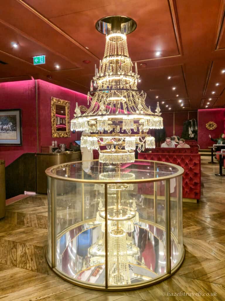 Large crystal chandelier displayed in a glass case inside the elegant Cafe Sacher, a Vienna coffee house with deep red walls and gold detailing.