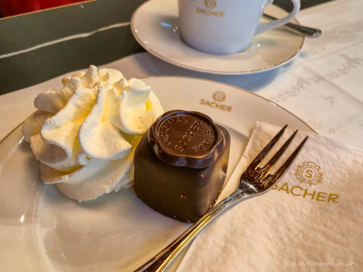 Chocolate-glazed Sachertorte slice topped with a round chocolate seal, served with a generous swirl of whipped cream and coffee in the background.