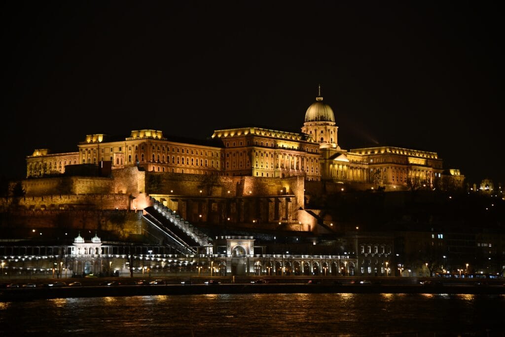 Buda Castle illuminated at night in Budapest, a large historic palace complex on a hillside with long rows of glowing windows and a central domed roof, rising above stone walls and stairways, with the dark Danube river in the foreground under a black night sky.