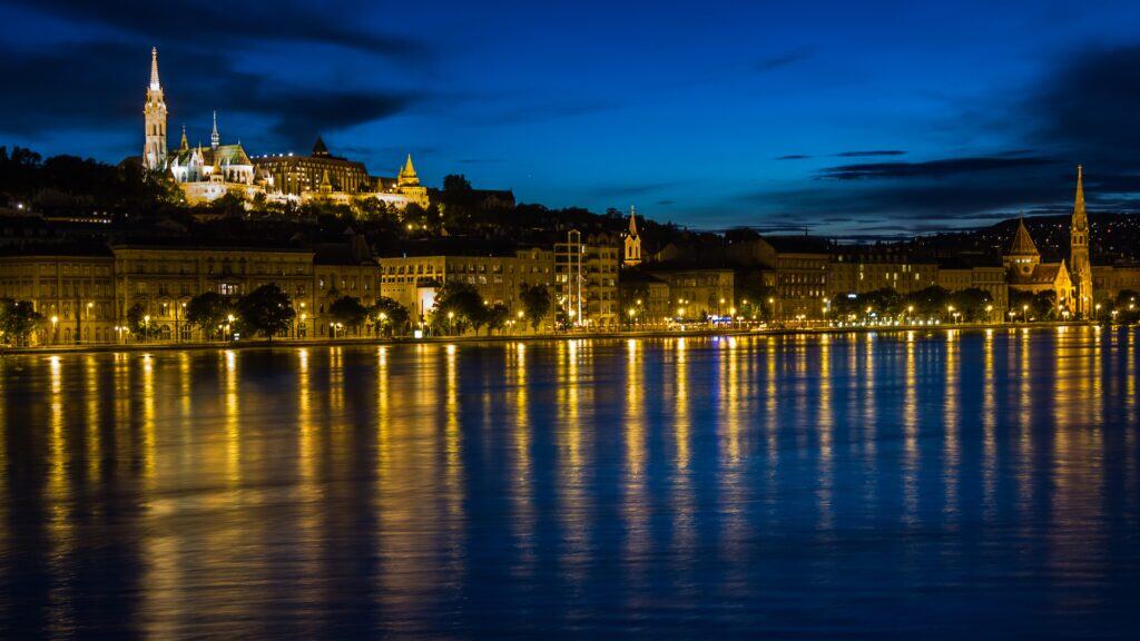 Night view across the Danube in Budapest, with a wide, calm river in the foreground reflecting long streaks of yellow and gold lights. On the far bank, rows of historic buildings line the waterfront, and on the hillside above them, Castle Hill rises with illuminated church towers and rooftops glowing against a deep blue evening sky.