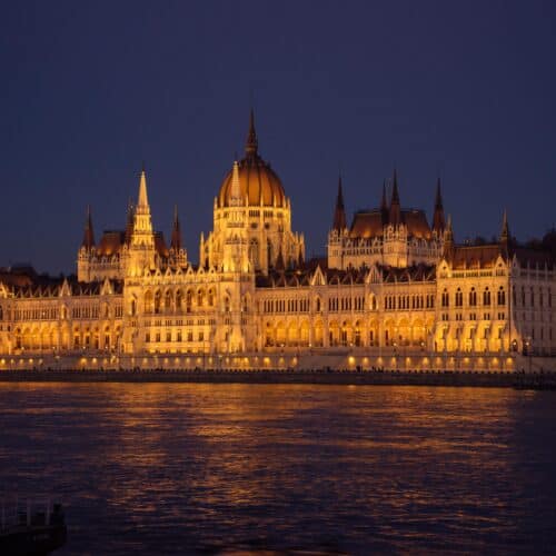 Hungarian Parliament building at night in Budapest, a very large stone building beside the Danube. It has a wide, symmetrical shape with a tall round dome in the centre, many narrow pointed towers, rows of arches, and bright yellow lights reflecting in the river below.