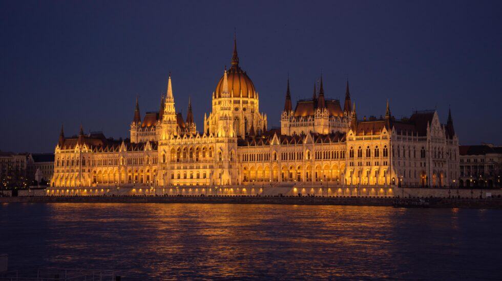 Hungarian Parliament building at night in Budapest, a very large stone building beside the Danube. It has a wide, symmetrical shape with a tall round dome in the centre, many narrow pointed towers, rows of arches, and bright yellow lights reflecting in the river below.