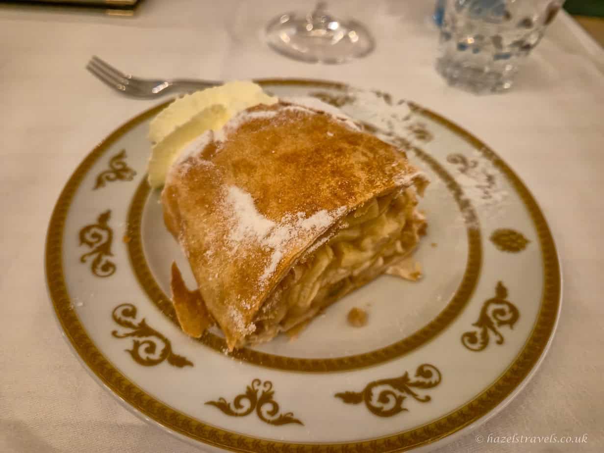Slice of Apfelstrudel dusted with icing sugar, showing layers of thin pastry and spiced apple filling on a decorative plate.