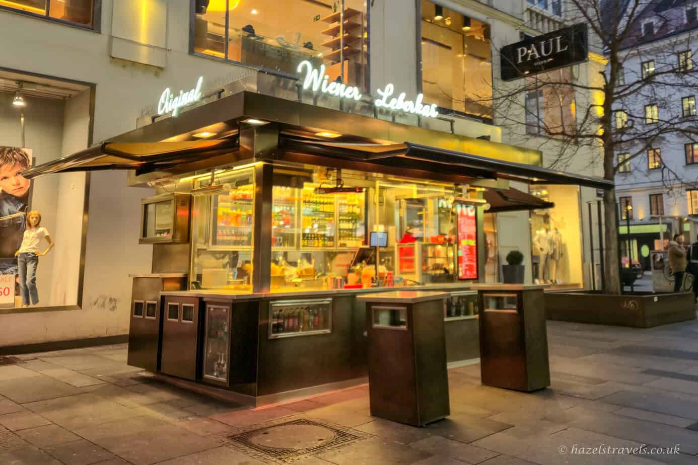 Traditional Viennese sausage stand (Würstelstand) lit up at dusk on a city street in Vienna.