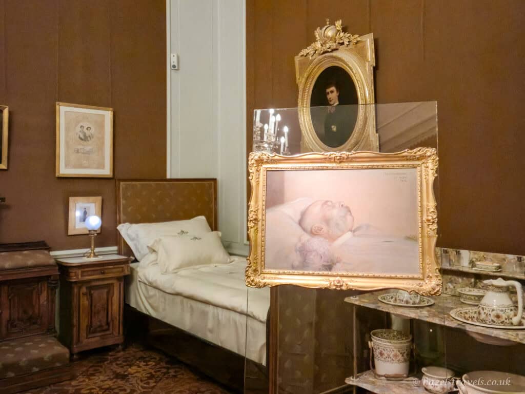 Inside a softly lit historic palace bedroom, a narrow single bed with crisp white linens and a cream pillow sits against warm brown walls. Beside it stands a dark wooden bedside table topped with a small glowing lamp. In front of the bed, behind a clear protective screen, hangs an ornate gold frame containing a pale portrait of an elderly man lying in repose. Above it, another gilded oval frame holds a smaller, darker portrait. To the right, a marble-topped washstand displays delicate patterned bowls and jugs, while framed photographs and drawings hang quietly on the surrounding walls, creating an intimate, preserved glimpse into a private royal room.