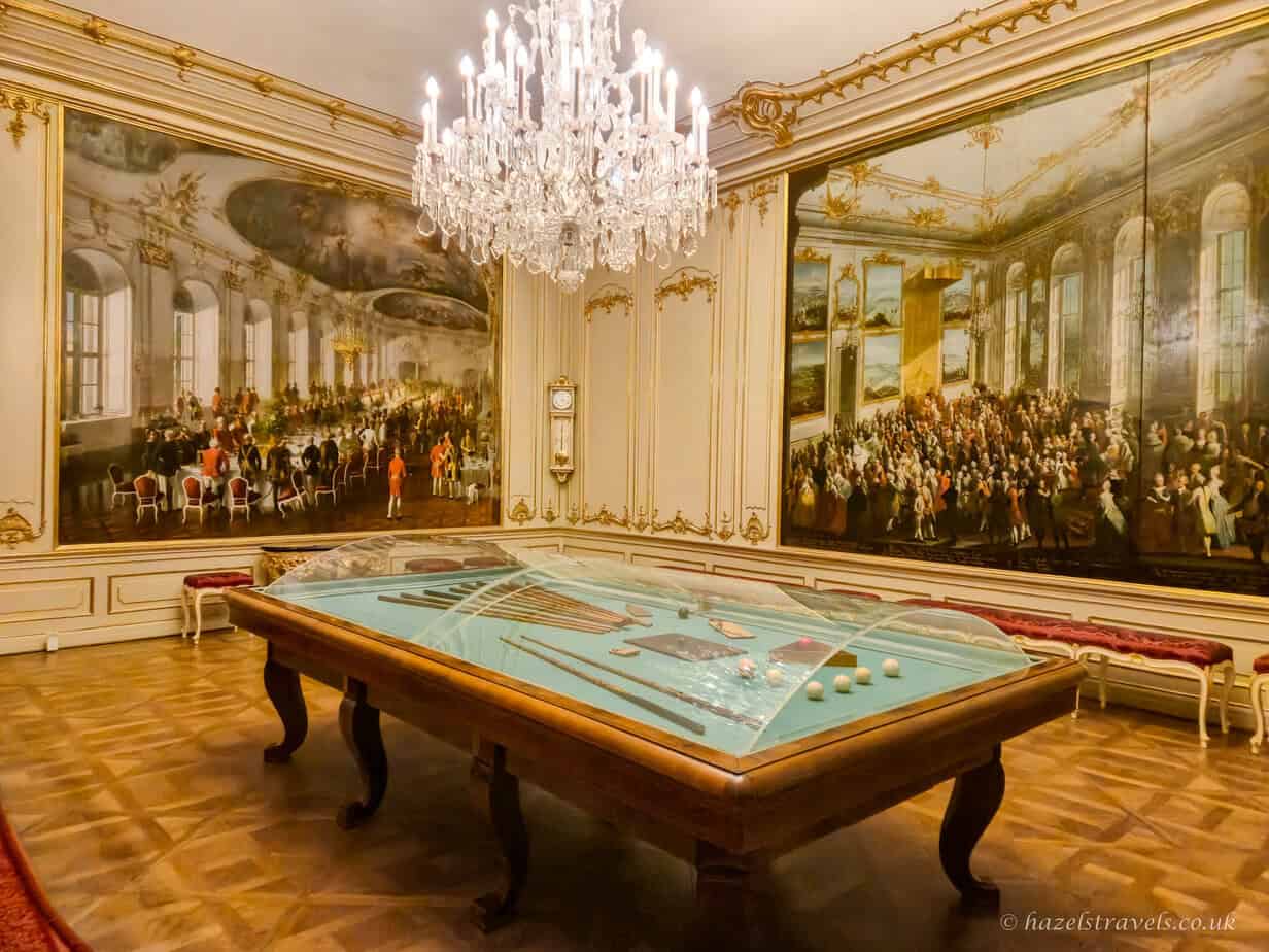 Large ceremonial hall inside Schönbrunn Palace featuring an enormous rectangular billiards table with a pale green felt surface and carved wooden legs. The polished parquet floor reflects light from a grand crystal chandelier overhead. Walls are richly decorated with white and gold Rococo mouldings, tall mirrors, and large historical paintings showing court scenes, creating a formal and opulent imperial atmosphere.