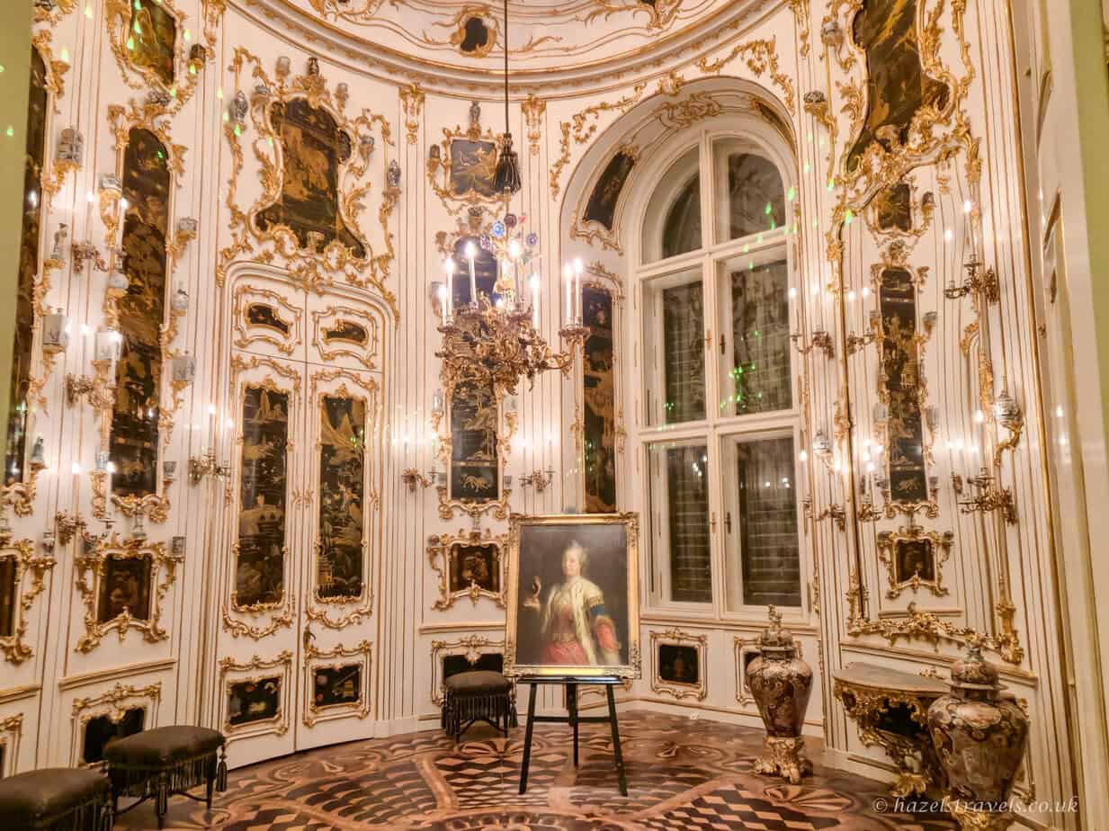 Ornate Rococo salon inside Schönbrunn Palace with curved white walls heavily embellished in gold leaf detailing. A tall arched window with multiple panes lets in soft light, framed by intricate gilded mouldings and crystal wall sconces. In the centre of the parquet floor stands a portrait of a young woman in a pale gown on an easel, surrounded by elegant antique chairs and small decorative tables, creating an intimate yet lavish imperial setting.