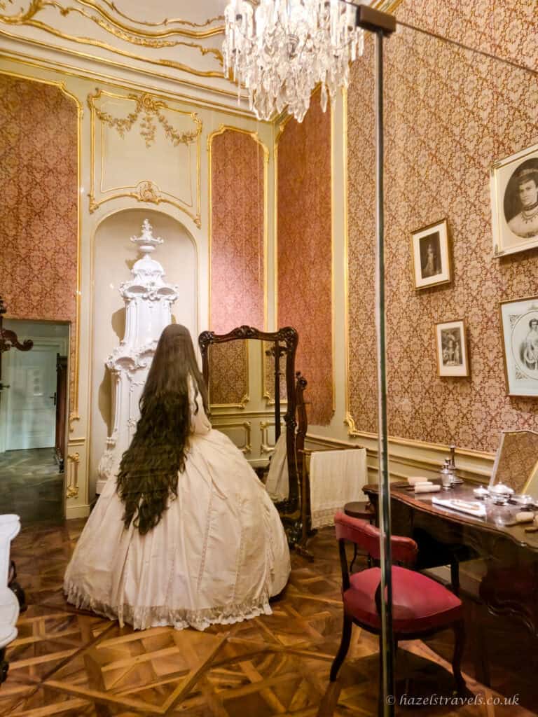 Recreated 19th-century bedroom scene inside Schönbrunn Palace, featuring a life-sized mannequin dressed in a voluminous ivory ball gown with an extremely long, dark wavy train cascading down her back. She stands before a tall, dark wooden cheval mirror on polished parquet flooring. The room is richly decorated with rose-patterned wallpaper framed by ornate gold mouldings, a crystal chandelier overhead, and a white ceramic tiled stove set into an arched alcove. To the right sits a dark wooden writing desk with silver toiletry items, framed black-and-white portraits on the wall above, and a red upholstered chair. A discreet glass barrier pole in the foreground indicates the display is part of the palace exhibition.