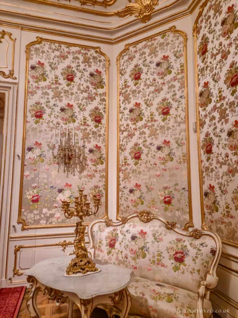 Cream-and-gold rococo palace wall panels filled with delicate floral wallpaper in soft pinks, reds, greens and lilacs, framed by ornate gilded mouldings. In front sits a small white-and-gold carved settee upholstered in matching floral fabric, beside a round marble-topped table with curved legs. A detailed gold candelabra with multiple candle arms stands on the table, completing the elegant, pastel-toned salon setting.