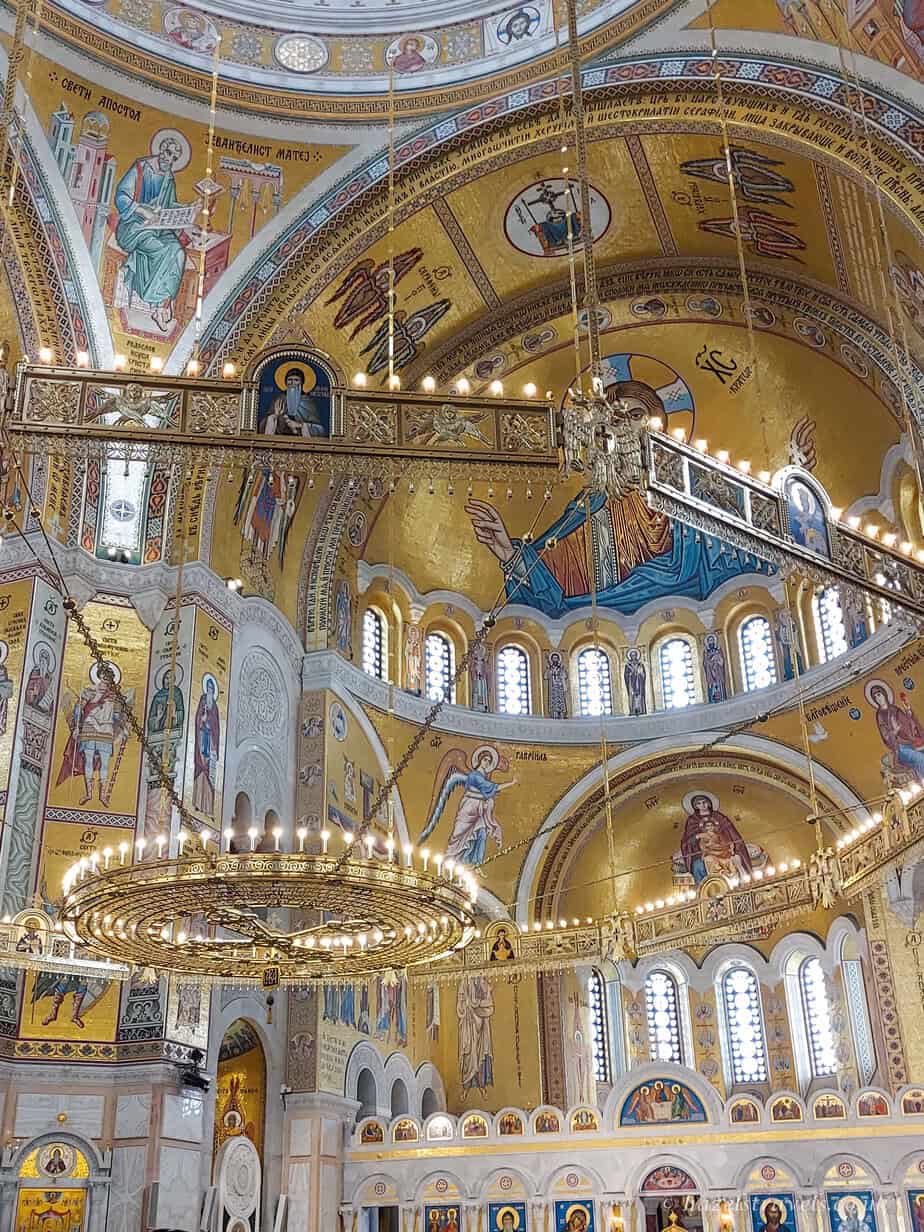 Interior of St Sava Temple in Belgrade, featuring dazzling golden mosaics, large circular chandeliers, arched windows, and detailed religious iconography on the domed ceilings and walls.
