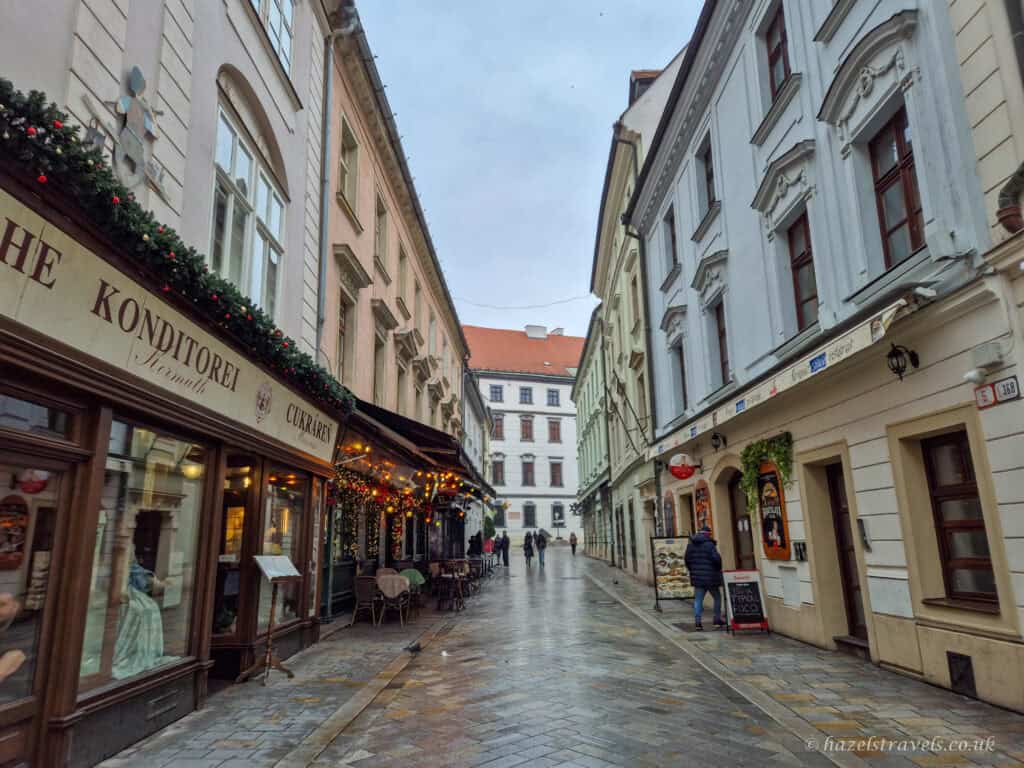 Narrow street in Bratislava Old Town with pastel historic buildings on both sides, a traditional café with festive garlands and outdoor tables on the left, wet cobbled pavement, and a few people walking under a grey winter sky.