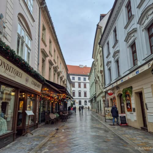 Narrow street in Bratislava Old Town with pastel historic buildings on both sides, a traditional café with festive garlands and outdoor tables on the left, wet cobbled pavement, and a few people walking under a grey winter sky.
