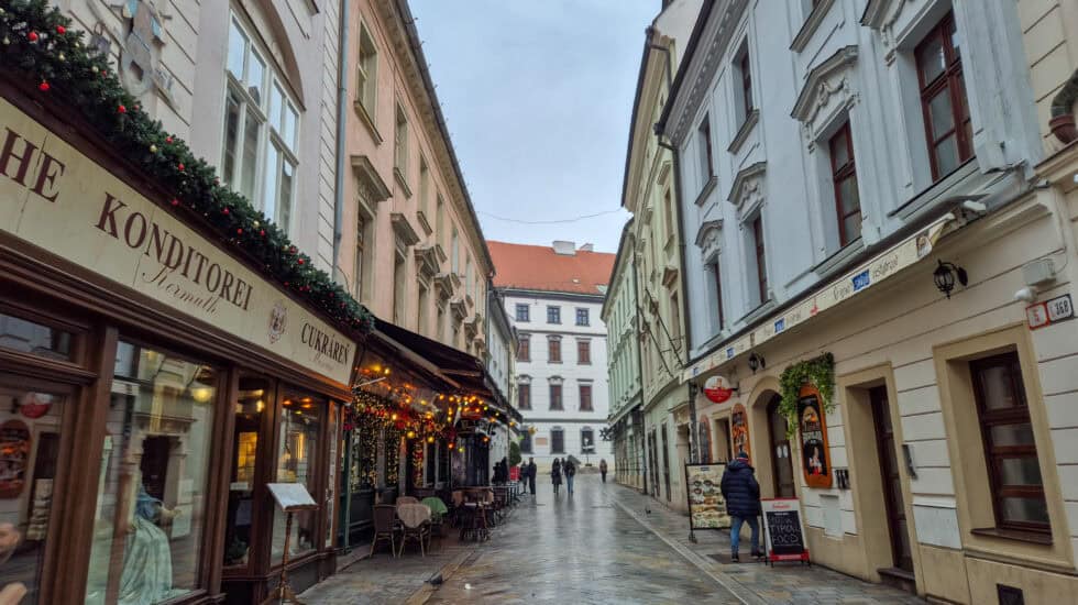Narrow street in Bratislava Old Town with pastel historic buildings on both sides, a traditional café with festive garlands and outdoor tables on the left, wet cobbled pavement, and a few people walking under a grey winter sky.