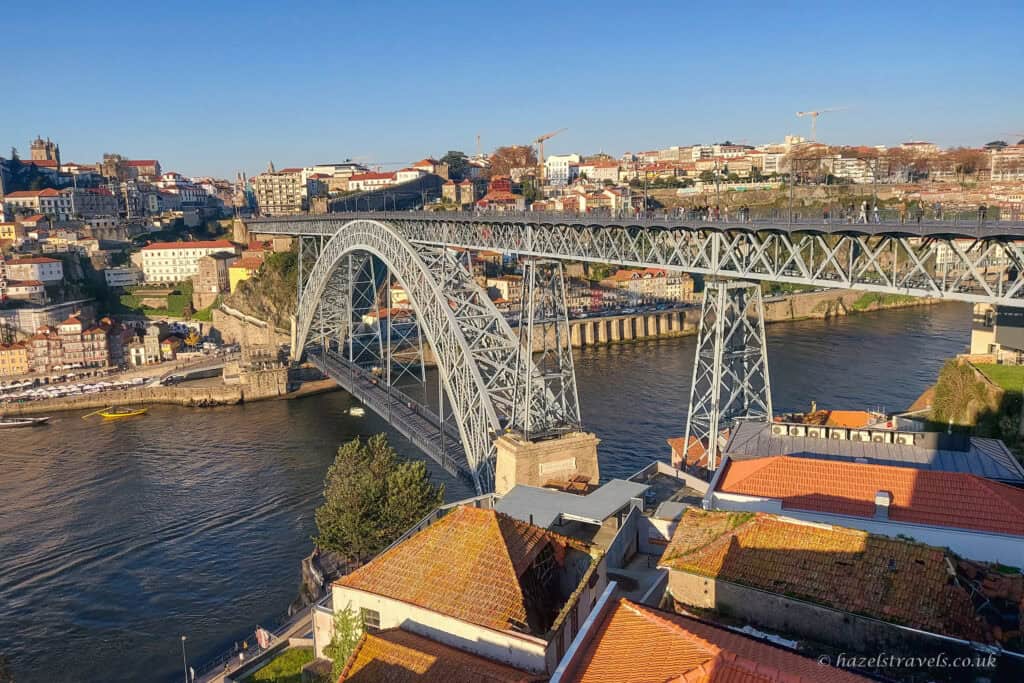 Elevated view of the Dom Luís I Bridge crossing the River Douro in Porto, with iron arches spanning the water, red-tiled rooftops in the foreground, and hillside buildings under a clear blue sky.