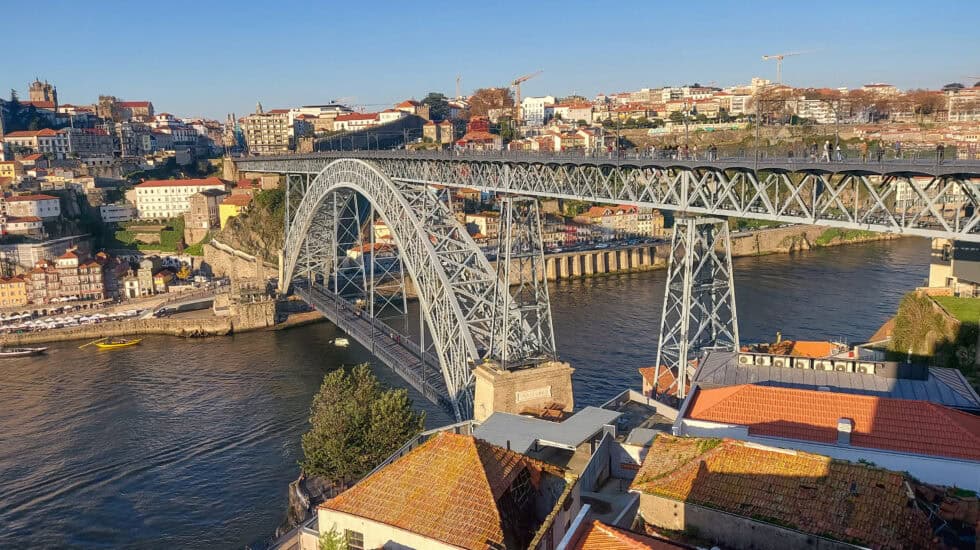 Elevated view of the Dom Luís I Bridge crossing the River Douro in Porto, with iron arches spanning the water, red-tiled rooftops in the foreground, and hillside buildings under a clear blue sky.