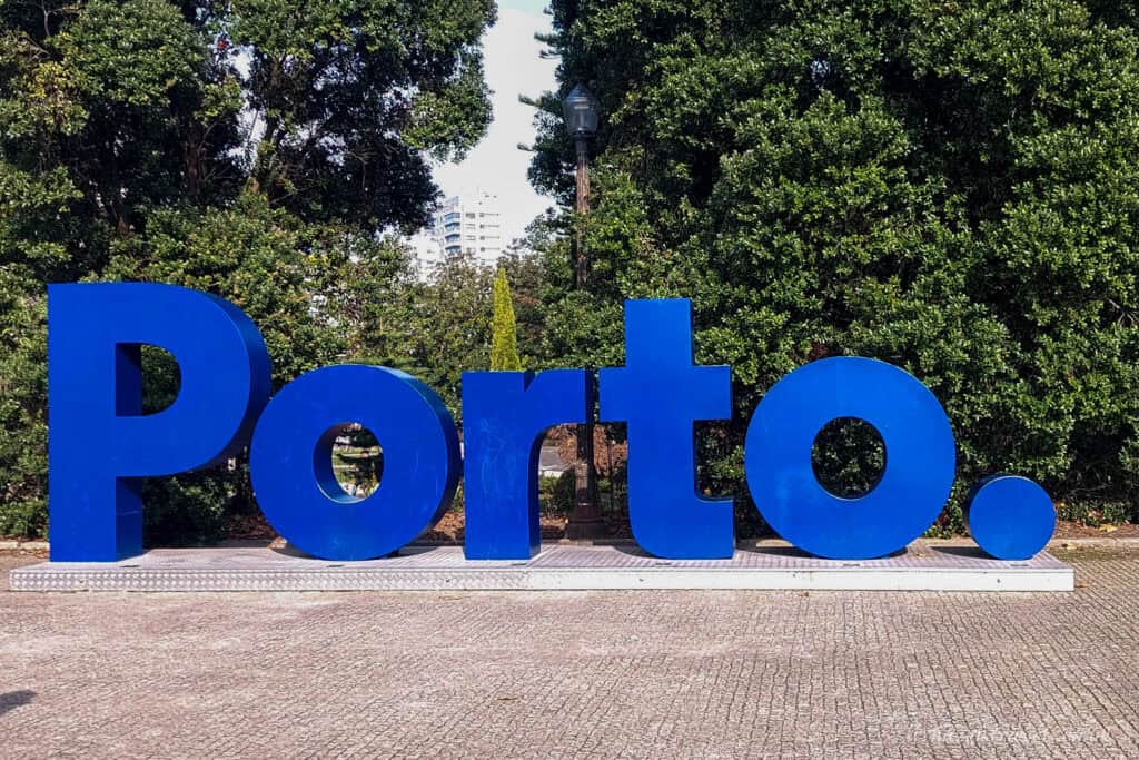 Large blue letters spelling “Porto.” displayed in a park setting, with trees and greenery behind the sign on a paved walkway.