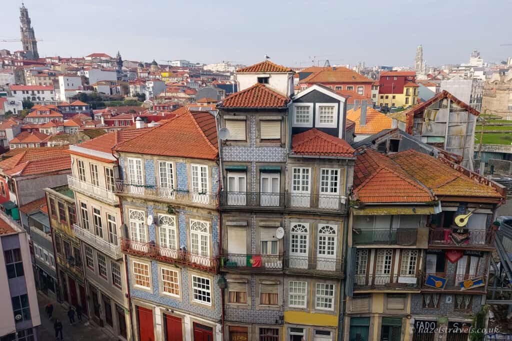 View over Porto’s historic centre with tightly packed buildings covered in blue-and-white azulejo tiles, red-tiled roofs, and wrought-iron balconies, stretching across the city under a pale sky.