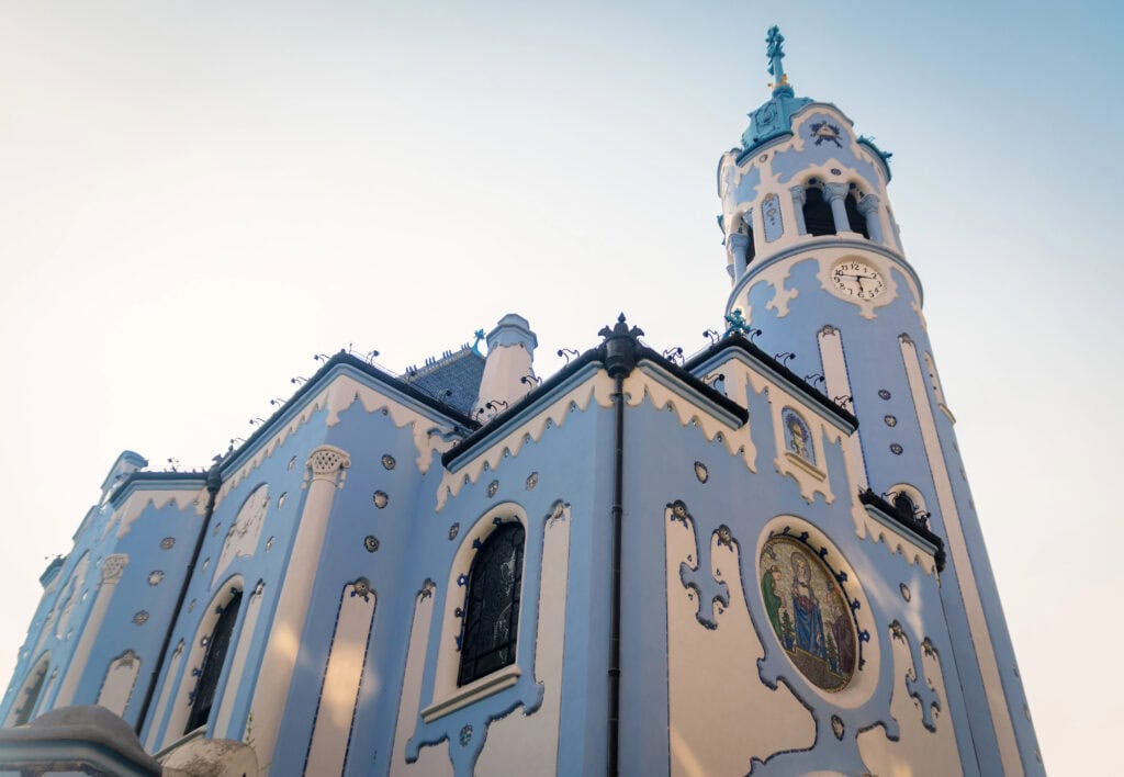 The Blue Church (Church of St Elizabeth) in Bratislava, painted pale blue with white decorative trim, rounded arches, mosaic details, and a tall clock tower rising against a clear light sky.