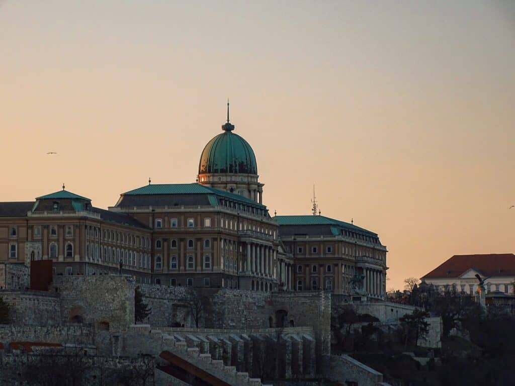 Buda Castle on Castle Hill in Budapest at dusk, with a large green copper dome and historic stone buildings silhouetted against a soft peach and pale blue evening sky.