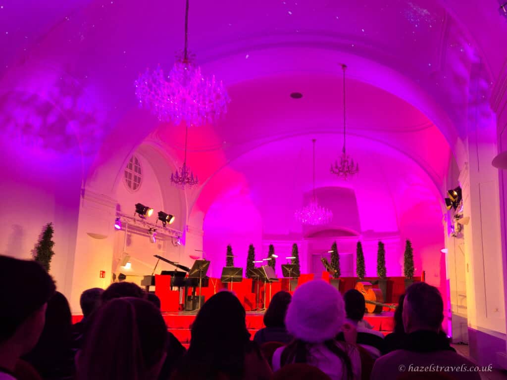 Audience seated beneath glowing pink chandeliers and vaulted ceiling before a Mozart concert at the Orangery, Schönbrunn Palace, Vienna