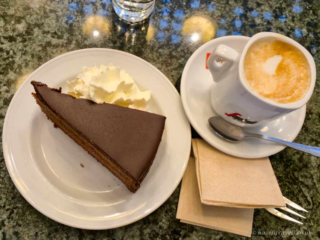 Slice of glossy chocolate cake topped with a swirl of whipped cream on a white plate, served alongside a frothy cappuccino in a white cup and saucer on a marble café table inside Schönbrunn Palace café.