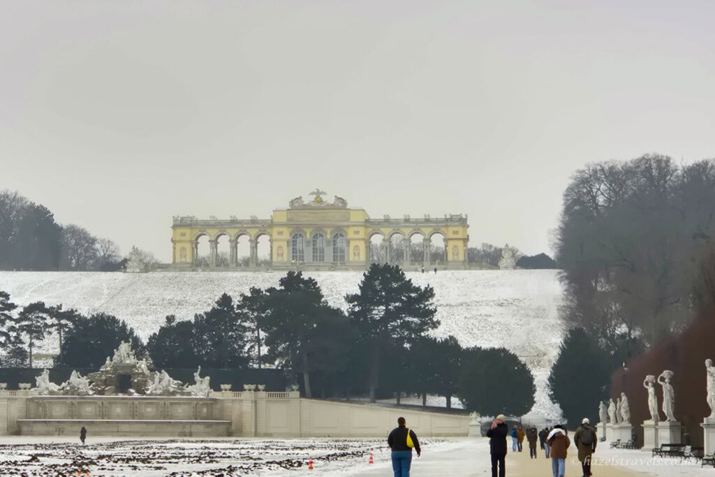 Gloriette at Schönbrunn Palace in Vienna, seen across snow-dusted gardens with Neptune Fountain in the foreground and visitors walking beneath a pale grey winter sky