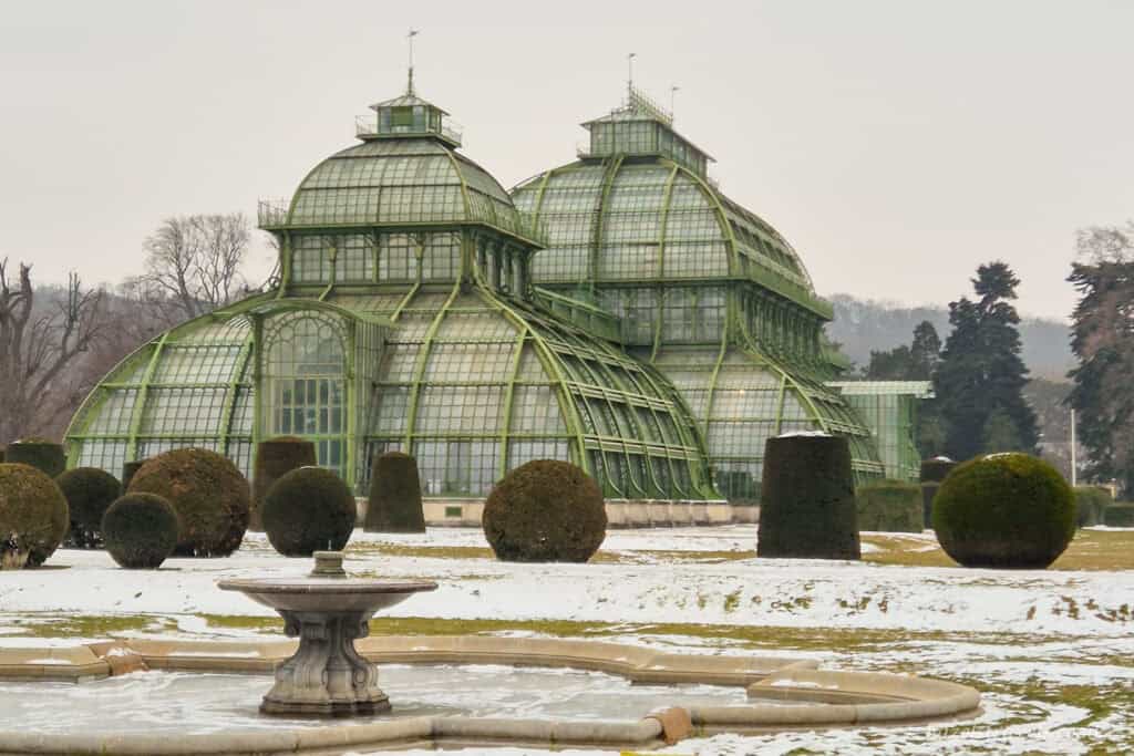 Palm House at Schönbrunn Palace in Vienna, a grand green glass greenhouse surrounded by snow-dusted gardens, clipped topiary shrubs and a stone fountain under a pale grey winter sky