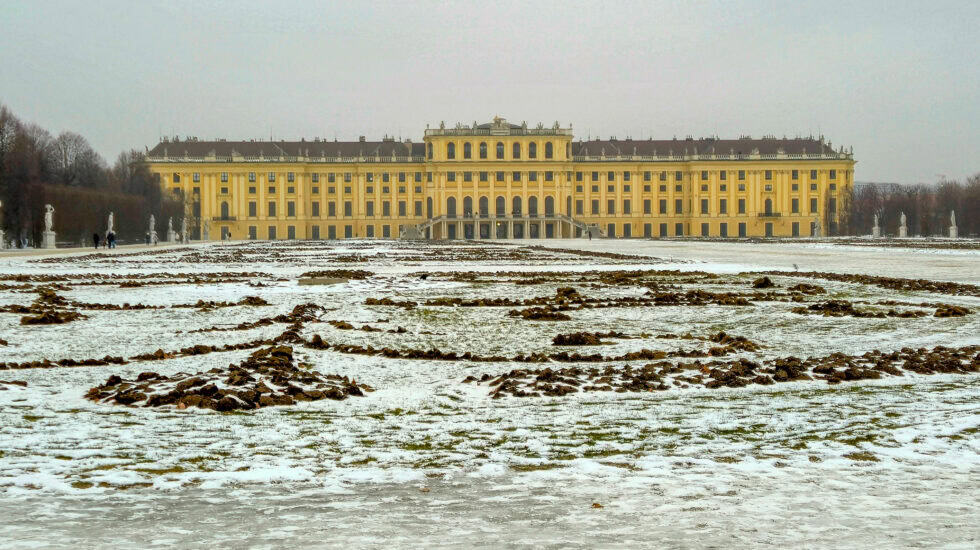 Schönbrunn Palace in Vienna viewed across snow-dusted formal gardens, with the yellow Baroque façade glowing beneath a flat grey winter sky