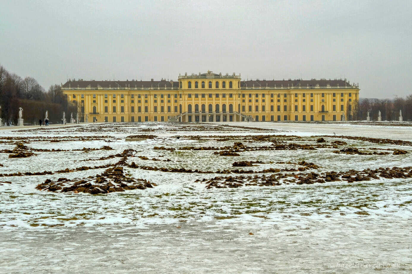 Schönbrunn Palace in Vienna viewed across snow-dusted formal gardens, with the yellow Baroque façade glowing beneath a flat grey winter sky