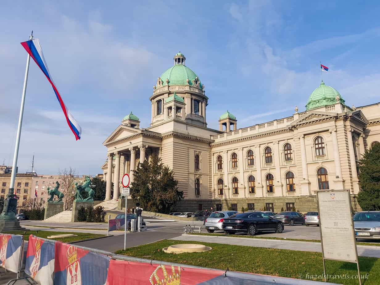The Serbian National Assembly building in Belgrade, with its green copper domes and grand neoclassical architecture, flying the Serbian flag under a bright blue sky, with a statue and parked cars in the foreground.