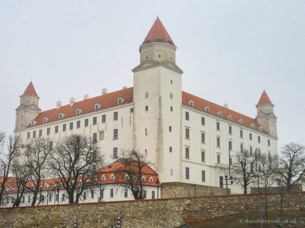 Bratislava Castle seen from below, with its large white rectangular walls, four corner towers topped with red-orange roofs, bare winter trees in the foreground, and a pale grey sky overhead.