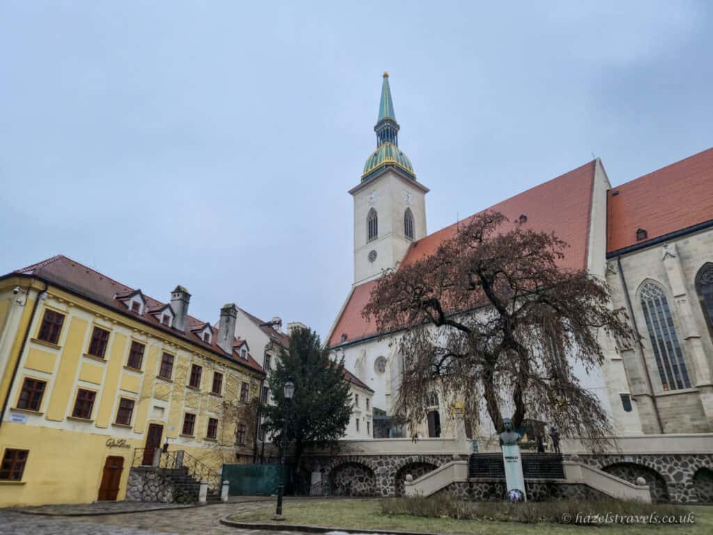 St Martin’s Cathedral in Bratislava, with its pale stone walls and tall tower topped by a green-and-gold spire, flanked by red-roofed historic buildings and a leafless tree in a quiet old town square under a cloudy sky.