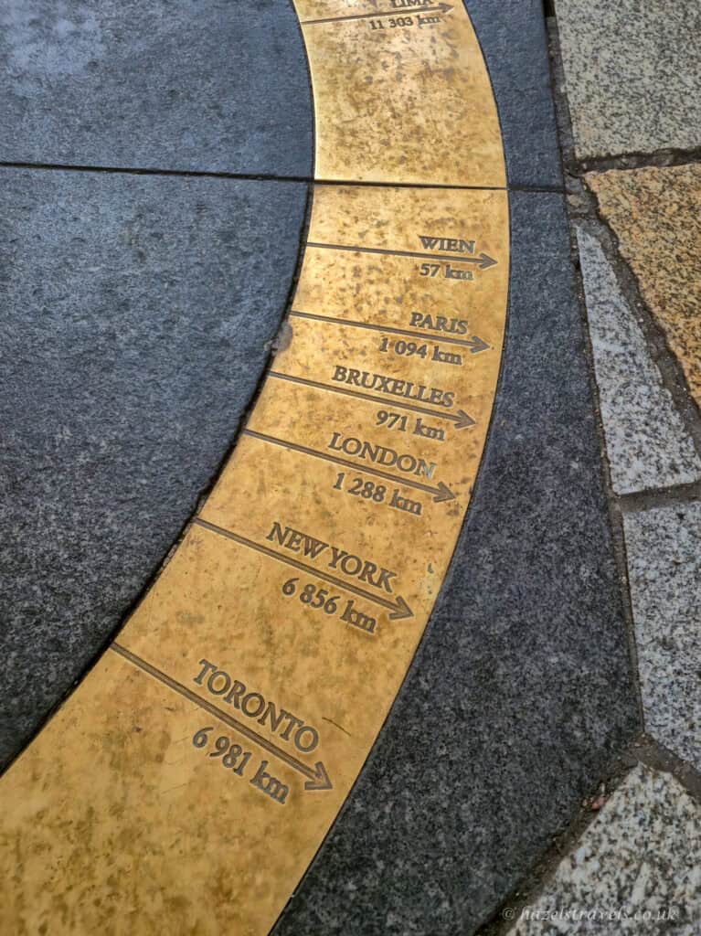 Curved brass inlay set into grey stone paving in Bratislava, engraved with arrows and distances to cities including Vienna, Paris, Brussels, London, New York, and Toronto, showing kilometres on a weathered gold-coloured surface.