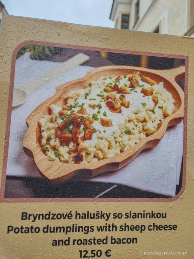 Menu sign in Bratislava showing a wooden bowl of bryndzové halušky, small potato dumplings topped with white sheep cheese, crispy golden-brown bacon pieces, and chopped green chives, photographed through glass dotted with rain droplets.