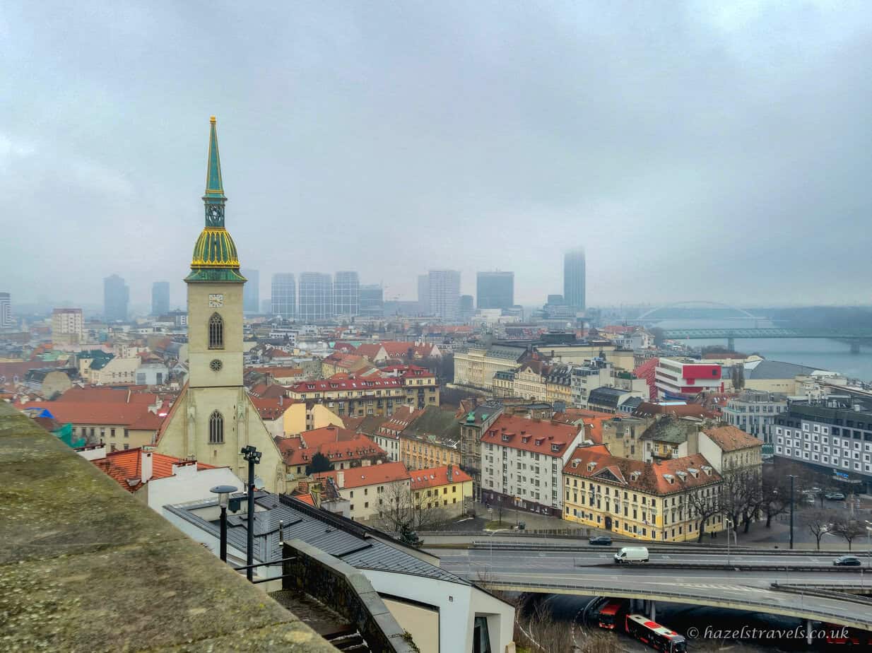 Elevated view over Bratislava Old Town with red and orange tiled rooftops, the tall tower and green-and-gold spire of St Martin’s Cathedral on the left, and modern high-rise buildings fading into mist in the distance under a grey sky.