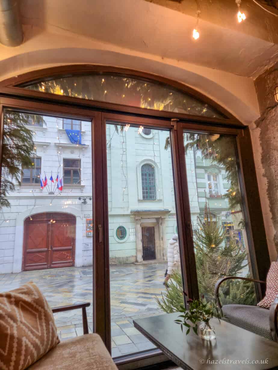 Cosy café interior in Bratislava with a curved ceiling, warm lighting, cushioned seating, and large arched windows looking out onto a quiet old town street with pale historic buildings outside.