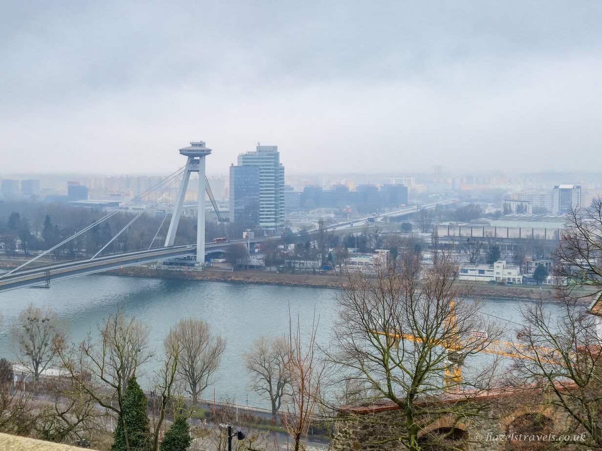View across the River Danube in Bratislava with the UFO Bridge and its tower in the distance, leafless winter trees in the foreground, muted blue-grey water, and a hazy skyline beneath an overcast sky.