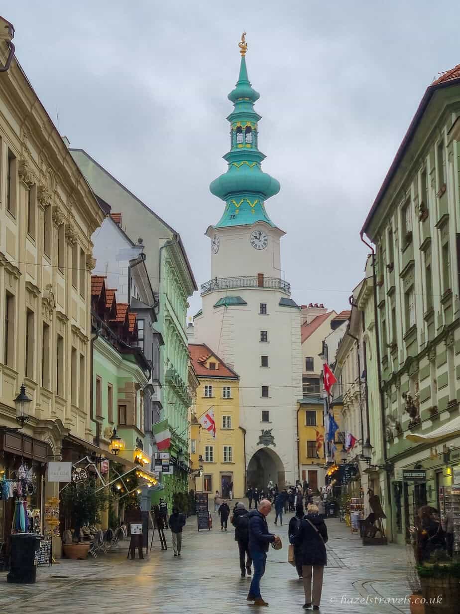 Pedestrian street in Bratislava Old Town lined with tall historic buildings, with people walking along the wet pavement toward Michael’s Gate, its green copper tower rising at the end of the street under a grey sky.