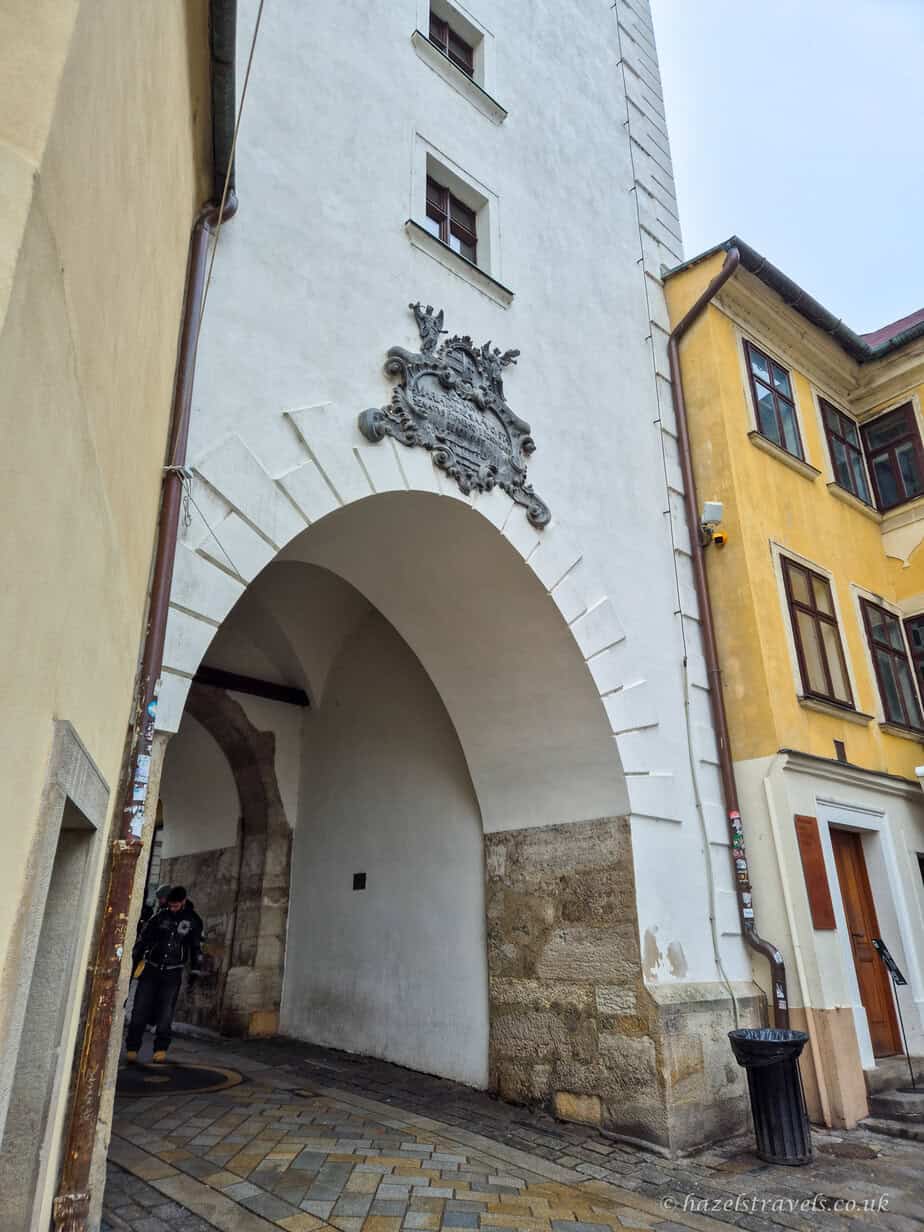 View through Michael’s Gate in Bratislava Old Town, showing the pale stone archway with decorative crest above, cobbled street below, and colourful yellow and cream historic buildings on either side.