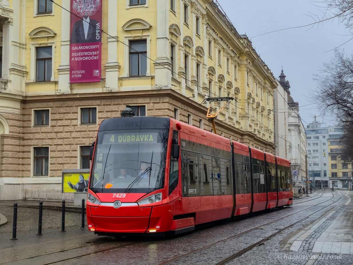 Red modern tram travelling through central Bratislava, passing pale yellow and cream historic buildings along a wet street, with overhead tram lines and a grey sky above.