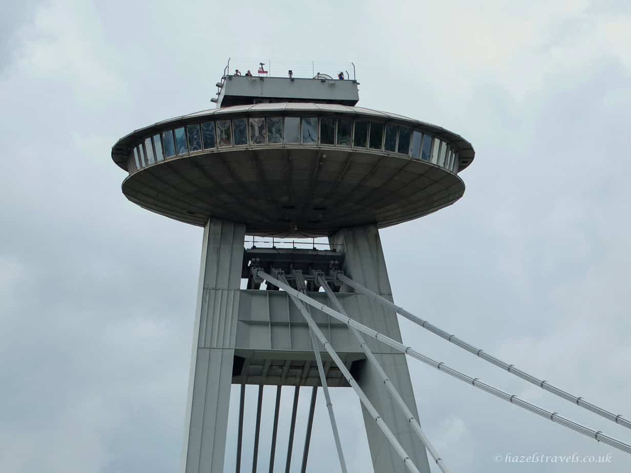 Upward view of the UFO Tower in Bratislava, with its circular saucer-shaped observation deck supported by tall grey pillars and suspension cables, set against a pale blue sky.
