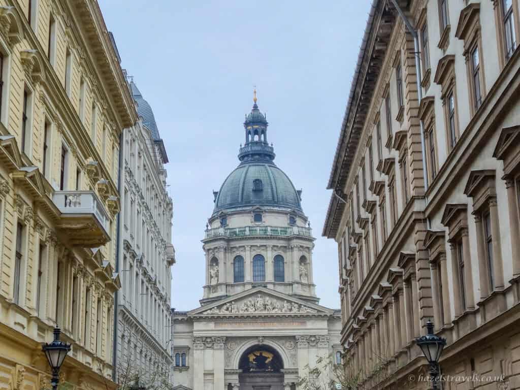 St. Stephen’s Basilica in Budapest seen in daylight, with its large green-domed roof framed by tall pale stone buildings on either side under a soft grey winter sky.
