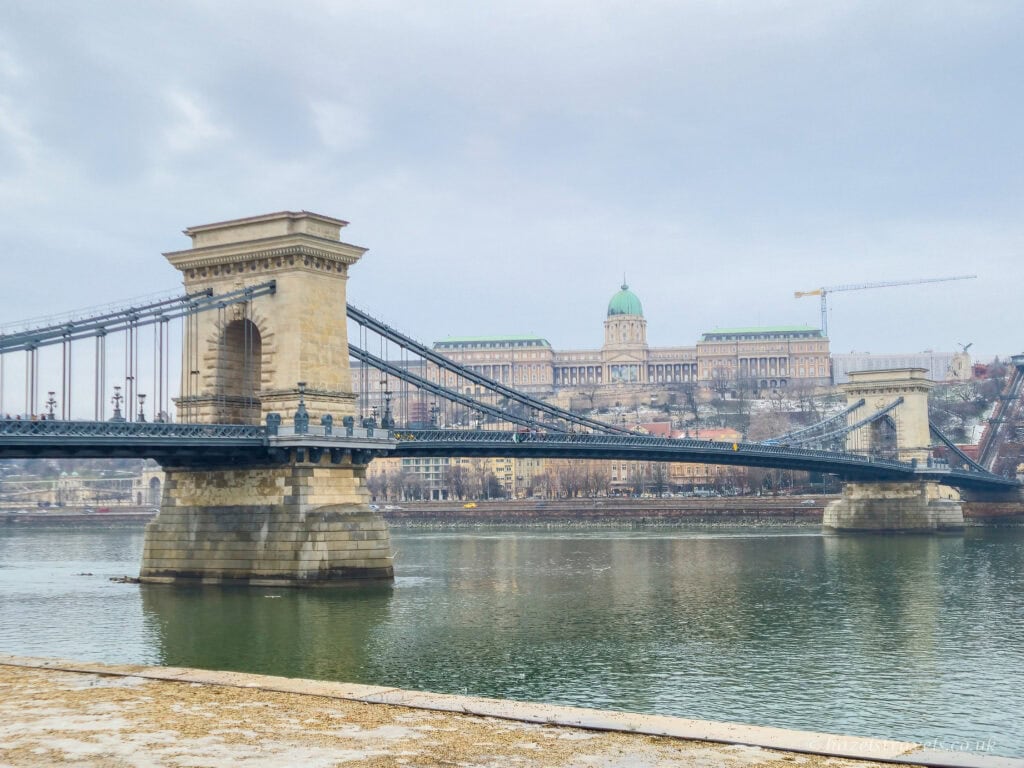 Chain Bridge in Budapest during winter daylight, with stone towers and suspension cables stretching across the Danube, Buda Castle visible on the hill in the background under a pale grey sky.