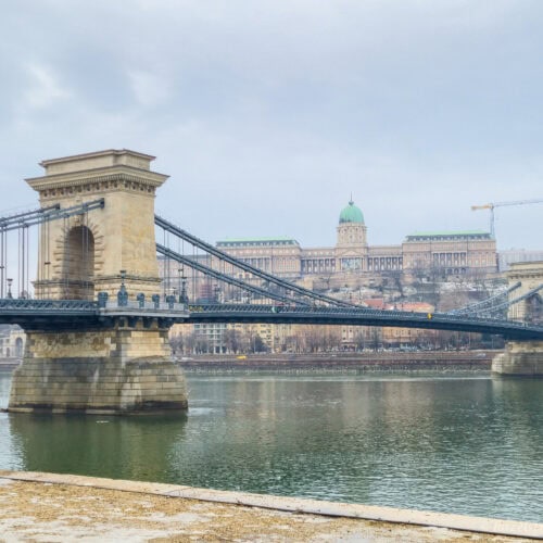 Chain Bridge in Budapest during winter daylight, with stone towers and suspension cables stretching across the Danube, Buda Castle visible on the hill in the background under a pale grey sky.