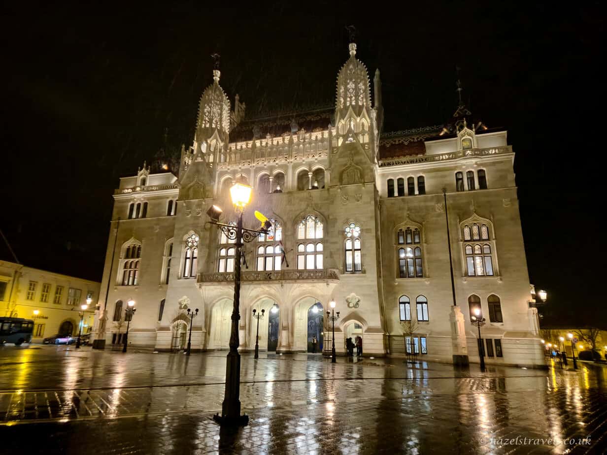 Ministry of Agriculture building in Budapest at night, with ornate neo-Gothic facade and twin spires lit in soft white light, wet stone square reflecting lamps and windows under a dark rainy sky.