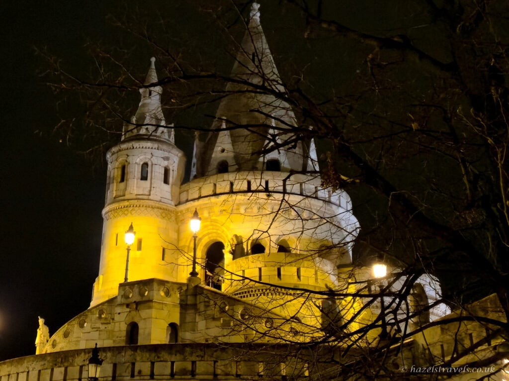 Fisherman’s Bastion in Budapest at night, with white stone towers and turrets glowing warm yellow under floodlights, bare tree branches silhouetted in the foreground against a dark winter sky.