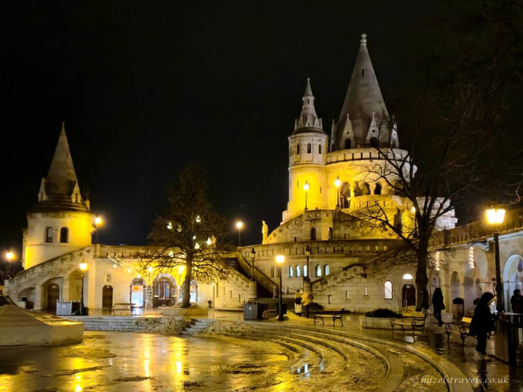 Fisherman’s Bastion in Budapest at night, with multiple white stone towers glowing yellow under floodlights, wet paving reflecting light in the foreground and a few people walking through the terrace area.